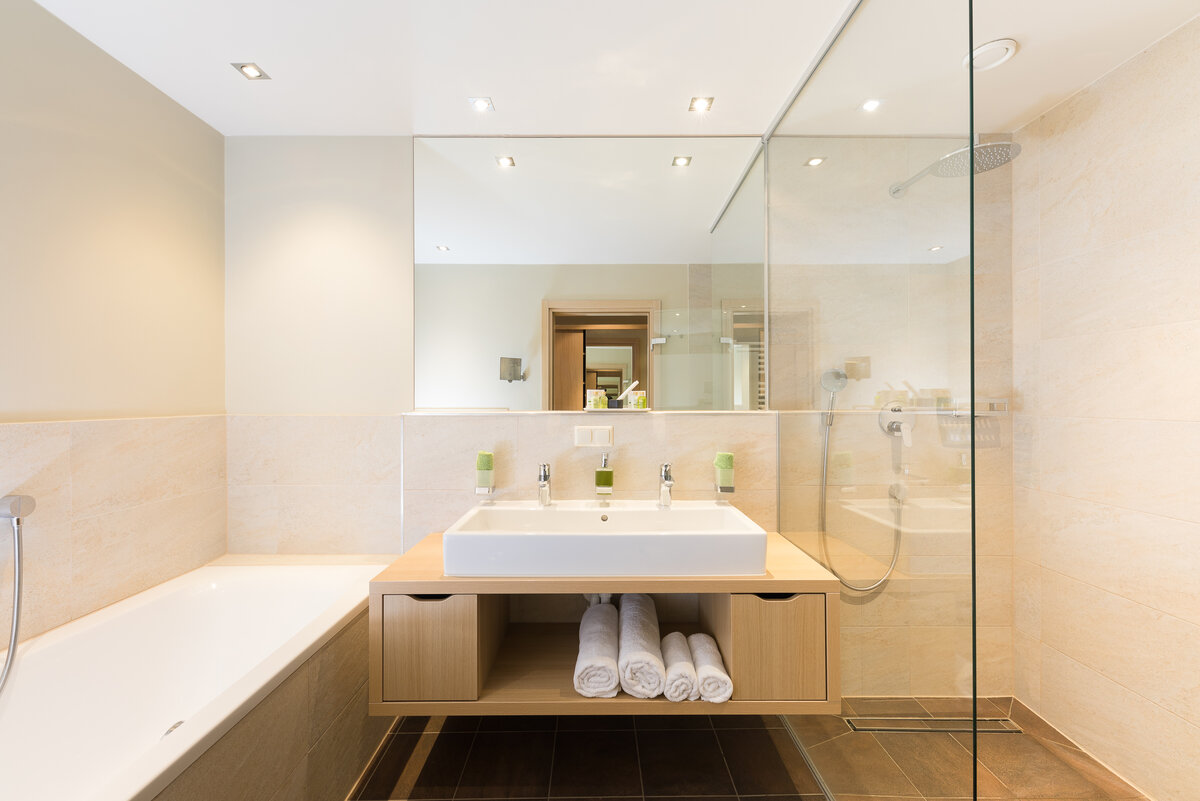 Modernes Badezimmer mit einer  Badewanne, einem Doppelwaschbecken auf einem Holzunterschrank, großen Spiegeln und einer begehbaren Dusche mit Glastrennwand.