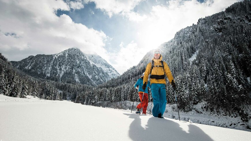 Zwei Personen wandern mit Schneeschuhen durch eine verschneite Winterlandschaft, umgeben von schneebedeckten Bergen und dichten Wäldern.